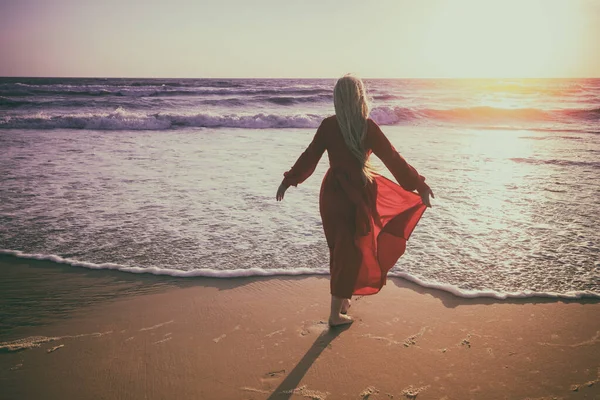 金色日落时的海景 海滩上的女人身穿红色飘扬衣服的年轻而快乐的女人沿着海滨散步 女孩看着神奇的落日 — 图库照片