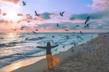Güneş doğarken deniz manzarası ve güzel bir gökyüzü. Elleri havada genç ve mutlu bir kadın turuncu bir elbiseyle kumsalda yürüyor. Martılar sahilde uçar.