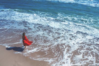 Kırmızı kanat çırpan elbiseli genç ve mutlu bir kadın deniz kıyısında yürüyor.