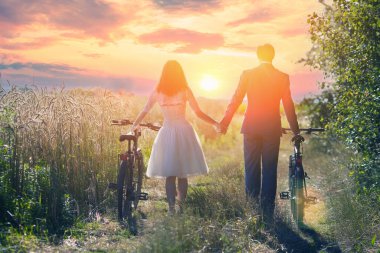 Bisikletli genç gelin ve damat gün batımında buğday tarlası boyunca el ele yürürler.
