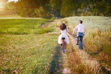Genç mutlu gelin ve damat çayırda bisiklete biniyor. Sırtları kameraya dönük el ele tutuşuyorlar.