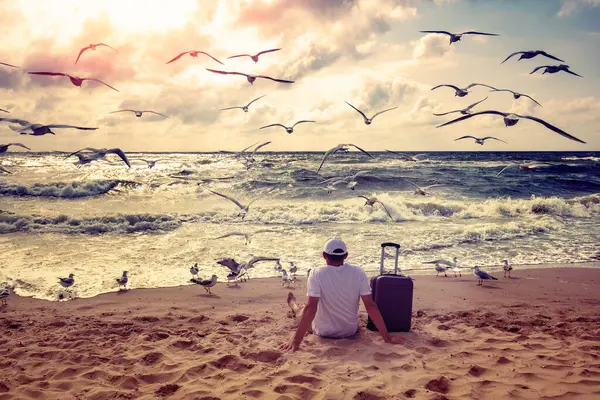 A human sits on the beach in summer with travel suitcase. The man looks at the sea. Seagulls fly over the beach. Travel, happy holiday, summertime concept