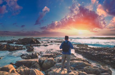 Akşamları deniz manzarası. Kaya sahili. Sahilde erkek turist sihirli gün batımını izliyor.