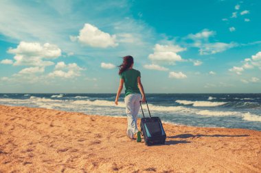 Güneşli bir günde deniz manzarası. Tatil konsepti, yaz zamanı. Kumsalda yürüyen seyahat çantalı genç bir kadın.