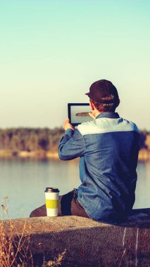 Bir adam akşamları güzel bir gölün beton setinde oturur. Bir adam tablet kullanarak gölün fotoğraflarını çekiyor. Dikey pankart
