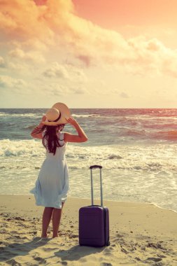 Hasır şapkalı, seyahat valizli genç ve mutlu bir kadın yazın sahilde yürüyor. Bir kadın hasır bir şapka tutuyor ve denize bakıyor. Seyahat, iyi tatiller, yaz konsepti. Dikey resim