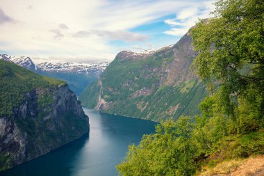 Bulutlu bir günde Geiranger fiyordunun manzarası. Ağaçların çerçevesiyle kayalık kıyılar. Norveç 'in güzel doğası