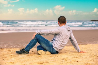 Genç adam sahilde oturuyor ve denize bakıyor.