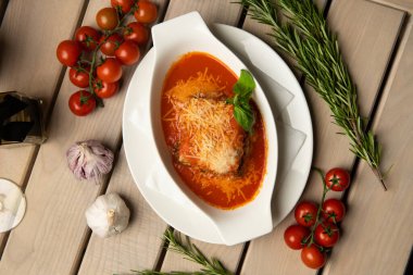 Beyaz tabakta fesleğen yapraklı lazanya. Klasik İtalyan yemeği işte. Masada domates ve sarımsak var. İtalyan mutfağı.