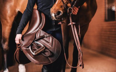 Unrecognizable Rider Standing Next to the Horse Inside Modern Red Brick Stable with Brown Leather Saddle in the Hand. Equestrian Lifestyle Theme. clipart