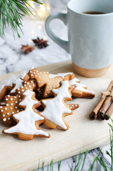 Festive cookies decorated with white icing on a wooden cutting board with a hot drink in a blue cup on a marble background with lights. Homemade ginger and cinnamon Christmas cookies. Vertical orientation.
