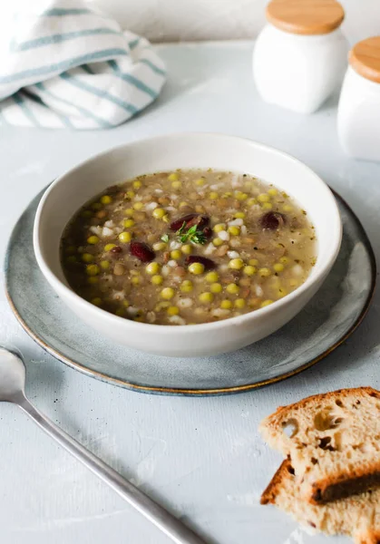 Bean soup in a ceramic gray bowl with a sprig of thyme and a piece of bread on a grey background. A mix of red beans, lentils and peas. The concept of vegetarian food. Vertical orientation.