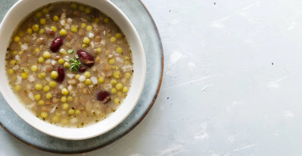 Bean soup in a ceramic gray bowl with a sprig of thyme and a piece of bread on a grey background. A mix of red beans, lentils and peas. The concept of vegetarian food. Horizontal orientation. Top view. Copy space. Banner