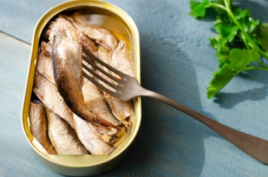 Konserve yemeğin yanında çatal üzerinde bir balık ve mavi ahşap bir masada maydanoz filizi. Konserve balık konsepti. Yatay yönelim.