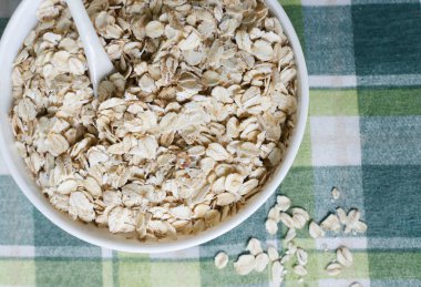 Yeşil kareli masa örtüsünde çay kaşığıyla beyaz bir kasede çiğ yulaf ezmesi. Sağlıklı kahvaltı kavramı. Vejetaryen ve vejetaryen. Yatay oryantasyon. Seçici odaklanma. Uzayı kopyala Üst görünüm