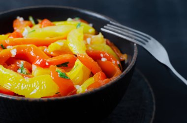 Kırmızı ve sarı kavrulmuş biber dilimleri, sarımsak ve maydanozlu salata siyah arka planda seramik kasede. Sağlıklı beslenme kavramı. Geleneksel İtalyan yemeği. Vejetaryen ve vejetaryen yemekleri.