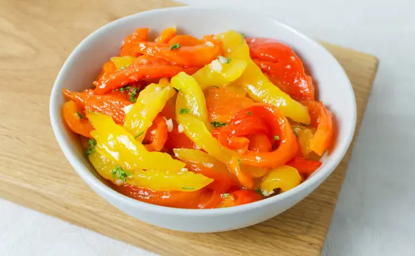 Dilimlenmiş kırmızı ve sarı kavrulmuş biber gri bir seramik kasede tahta bir kesme tahtasının üzerinde. Geleneksel İtalyan yemeği kavramı. Vejetaryen ve vejetaryen yemekleri. Yatay yönelim.