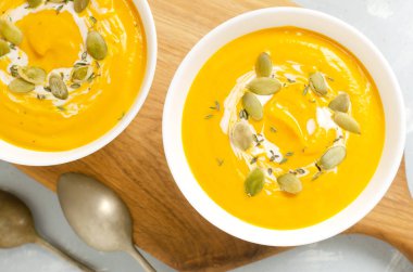 Gri arka planda tahta kesim tahtasının üzerinde kurumuş tohumlar ve kekik yapraklarıyla iki kase balkabağı kreması çorbası. Sağlıklı beslenme kavramı. Vejetaryen ve vejetaryen yemekleri. Üst görünüm