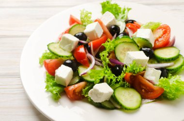 Beyaz ahşap bir masada salatalık, domates, kırmızı soğan, feta peyniri, marul ve siyah zeytinli klasik Yunan salatası. Geleneksel yemek kavramı. Sağlıklı yiyecekler. Basit malzemeler..