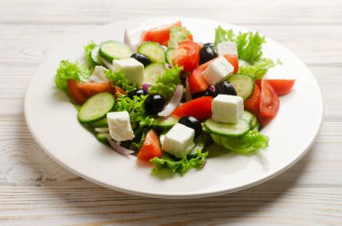 Beyaz ahşap bir masada salatalık, domates, kırmızı soğan, feta peyniri, marul ve siyah zeytinli klasik Yunan salatası. Geleneksel yemek kavramı. Sağlıklı yiyecekler. Basit malzemeler..