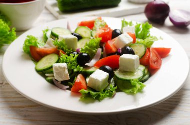 Beyaz ahşap bir masada salatalık, domates, kırmızı soğan, feta peyniri, marul ve siyah zeytinli klasik Yunan salatası. Geleneksel yemek kavramı. Sağlıklı yiyecekler. Basit malzemeler..