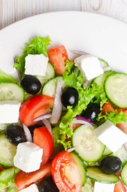 Beyaz ahşap bir masada salatalık, domates, kırmızı soğan, feta peyniri, marul ve siyah zeytinli klasik Yunan salatası. Geleneksel yemek kavramı. Basit malzemeler. Üst görünüm.