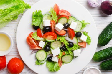 Beyaz ahşap bir masada salatalık, domates, kırmızı soğan, feta peyniri, marul ve siyah zeytinli klasik Yunan salatası. Geleneksel yemek kavramı. Basit malzemeler. Üst görünüm.