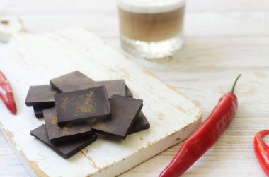 Masada bir bardak kapuçino ve beyaz ahşap kesme tahtasının üzerinde üç kırmızı biberli ekstra koyu çikolata parçaları. Trend gıda konsepti. Seçici odaklanma. Yatay yönelim.