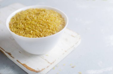 Seramik beyaz bir kasede gri arka planda beyaz bir kesme tahtasının üzerinde çiğ bulgur taneleri. Sağlıklı beslenme konsepti. Vejetaryen ve vejetaryen yemekleri. Kırsal tarzda. Yatay oryantasyon. Boşluğu kopyala