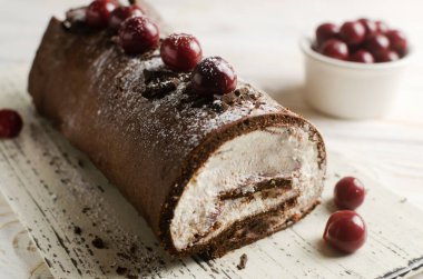 Black Forest roll cake with chocolate and cherries on a white plate. Homemade delicious pastries with delicate cream. Retro recipe concept. Horizontal orientation. Selective focus.