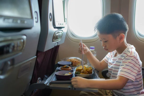 Asian Child in airplane window seat. Kids flight meal. Children fly. Special inflight menu, food and drink for baby and kid. Boy eating healthy lunch in airplane. Travel and family vacation.