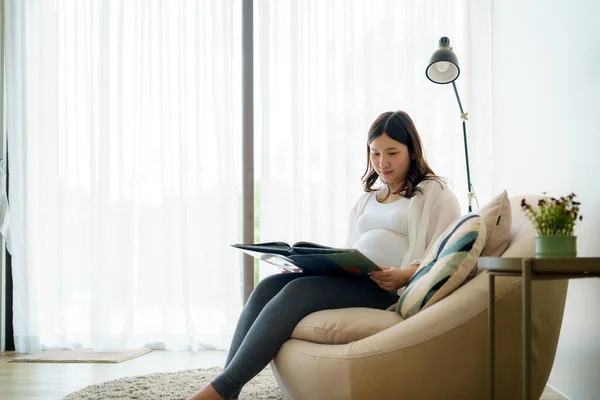Asian Pregnant woman read a book for baby in belly. Mom feeling happy smiling positive and peaceful while take care child lying on sofa in living room at home concept.