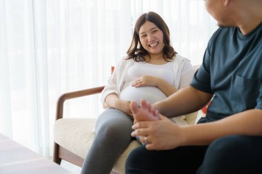 Asian husband does massage of feet of the pregnant wife in living room at house. Happy pregnant family.