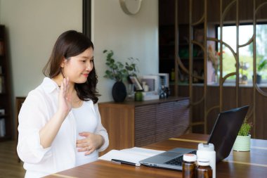 Asyalı hamile bir kadın elinde ilaç şişesiyle evdeki oturma odasında doktorla internet üzerinden iletişim kuruyor. Teletıp, hamilelik, bebek ve sağlık sigortası konsepti