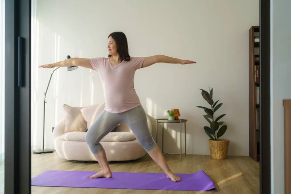 Full length healthy 6 months pregnant calm Asian woman meditating or doing yoga exercise at home. Relaxation and stretching.