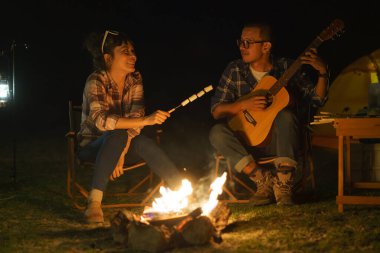 Asian woman toasting marshmallows while her boyfriend plays guitar At the campfire where they set up their tents to camp by the lake at night.