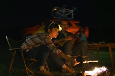 Asian woman toasting marshmallows while her boyfriend plays guitar At the campfire where they set up their tents to camp by the lake at night.