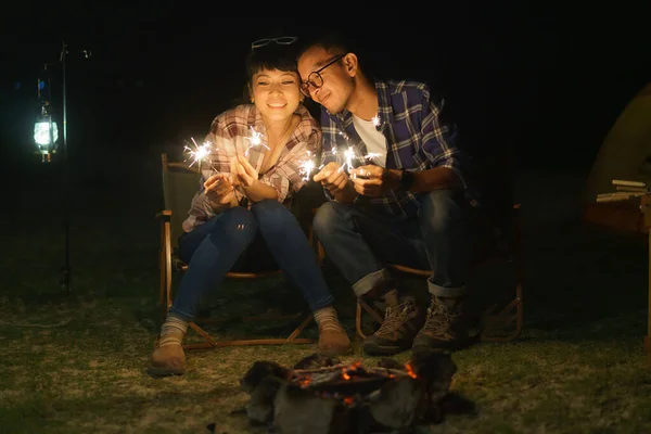 Asian couple is lighting sparkler fire at a campfire where they set up a tent to camp by the lake at night.