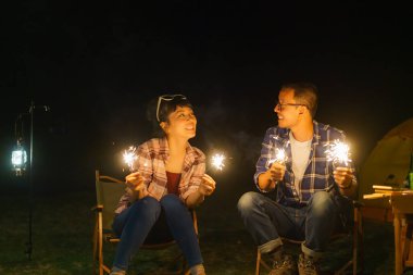 Asian couple is lighting sparkler fire at a campfire where they set up a tent to camp by the lake at night.