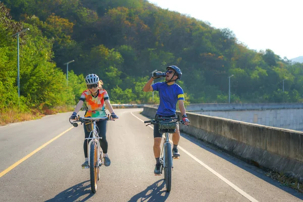 疲れたアジア人のサイクリストは 朝に湖の周りで運動するために一緒に乗っている間 ボトルから水を飲みます — ストック写真