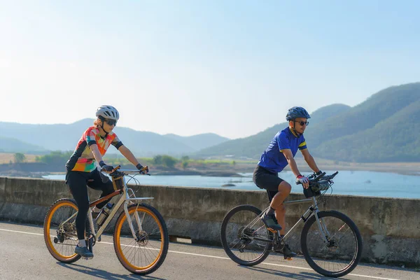 背景に美しい山の景色と朝の湖の周りの運動のために一緒に乗るアジアのサイクリストカップル — ストック写真