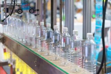 Görüntü, şişeleme fabrikasındaki bir taşıma bandında bir sıra temiz plastik su şişesini gösteriyor. Arka plandaki şişelere ve endüstriyel makinelere odaklanıyor.