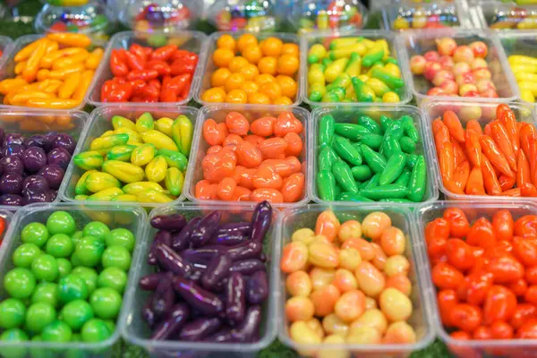 An assortment of Fruit Shaped Mung Beans vibrant Thai desserts, designed to resemble miniature fruits and vegetables, displayed in clear containers at a market