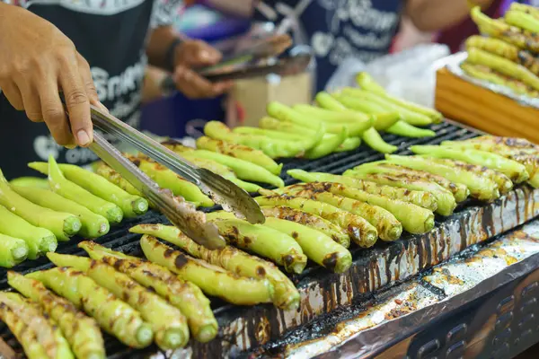 A street food vendor expertly grills a batch of fresh green chilies, getting them charred and ready to be served as a spicy side dish