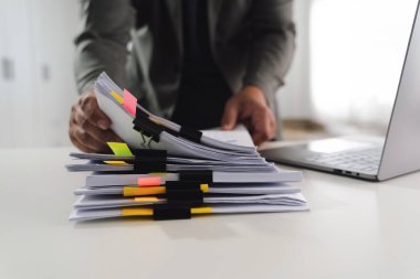 Dizüstü bilgisayarın yanında renkli not masası olan belgeleri düzenleyen, yoğun çalışmayı veya proje yönetimini gösteren bir belgeyi imzalayan bir kalem