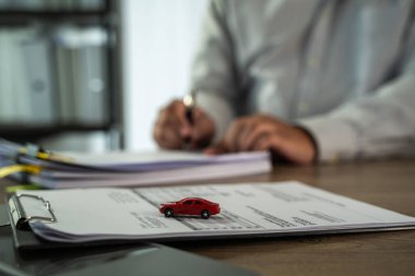 Evrak sigortası yapan gri gömlekli kişi evrakların üzerine küçük kırmızı bir oyuncak araba yerleştirip araba sigortası yaptırıyor.