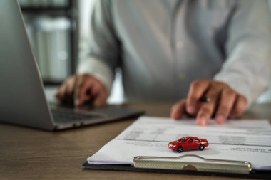 Evrak sigortası uzmanı, dizüstü bilgisayarla masa başında çalışıyor. Küçük kırmızı araba modelli bir belgeye işaret ediyor.