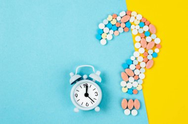 A question mark from laid out of medicine pills with Alarm Clock on blue background.