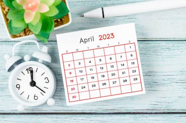 April 2023 Monthly calendar year and alarm clock with pen on blue wooden background. clipart
