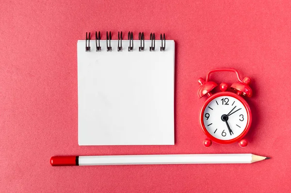 Top view of open empty spiral notebook and alarm clock with pen on red background.